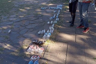Two men standing in a park, looking down at a collection of plastic bottles. The bottles have been sorted by type and laid in groups on the ground.