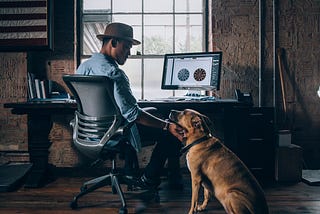 A designer patting his brown dog that is seated next to his desk.