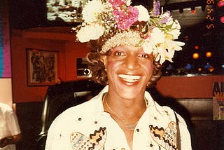 A photo of Marsha P. Johnson in a bar. She is wearing a large headpiece of flowers and smiling hugely at the camera.