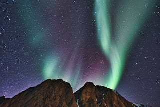 Green and pink aurorae on a black and purple sky dotted with white stars. A hill is in the foreground in the lower quarter of the image.