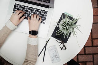 A picture describing a woman working on a laptop in a nice environment. On a table, there are two pencils, a cell phone, specs,  a little plant, and a laptop.