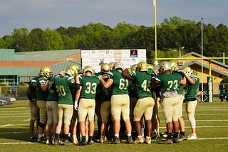 A team of high school football players in green and gold uniforms huddle up around the 50 yard line.