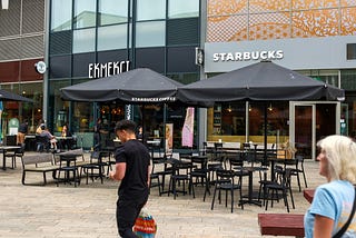 A city square lined with cafes, where only two people are present: an old lady and a young man walking.