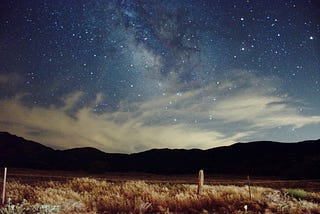 A fenced in field in front of mountains with a large view of the stars and the night sky.