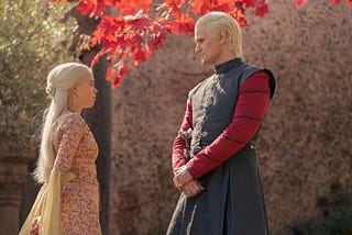 Rhaenyra and Daemon Targaryen standing beneath a tree with red leaves visible above their heads.