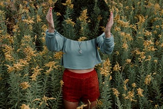 A woman in the middle of a flower field holds a circular mirror to the camera, reflecting the flowers and hiding her face.