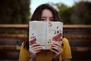 White woman with auburn hair and wearing a yellow shirt holds an open copy of The Handmaid’s Tale in front of her face.