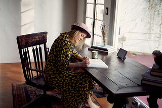 White woman wiht blond hair and a mauve hat writing on a notebook on a wooden desk and chair in a courtyard.