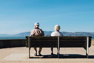 An old couple sitting on a bench staring out in the open looking at the mountains.