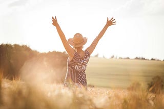 A person wearing a sunhat and dress with arms outstretched upwards in a sunlit field, embodying the feeking of freedom