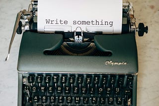 A black type writer with a sheet of paper that states “write something”.