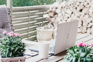 A laptop computer sits on a wooden table outside surrounded by plants. A stack of firewood is shown in the background.