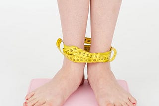 Image showing someone’s legs, standing on a weighing machine with an inch tape wrapped around their ankles.