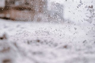 A snow-spackled window shows a blurry display of snow carpeting the ground and a large brown building in the far background.