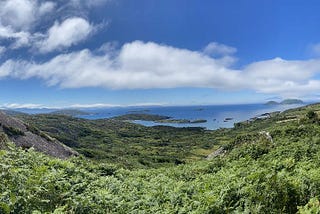 Photo from a hilltop in Derrynane, County Kerry, Ireland, with lush greenery in the foreground and small islands dotting the ocean in the distance. The sky is blue with puffy clouds.