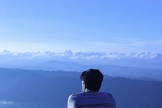 A young man sits on a hilltop, facing away and looking out over a series of hills and valleys.