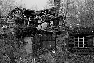 A black and white picture of an old, decaying home or barn. Rafters are exposed and toppling over. The building is barely still standing.