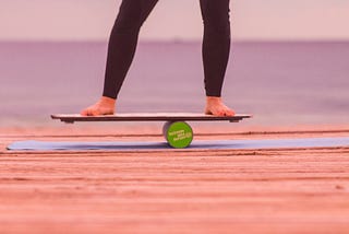 An image of legs from the knees down wearing black leggings and bare feet balancing on a board that is on a round green cylinder.
