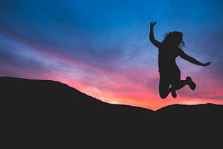 silhouette of female leaping joyfully in the air against a sunset sky and mountains in the background