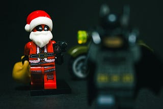 A Lego Deadpool with a Santa Claus beard, hat, and sack of presents walking up to a out of zoom Batman.