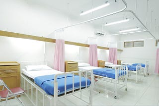a line of hospital beds in a white ward. the bed sheets are blue and the curtain divider between each bed is pink