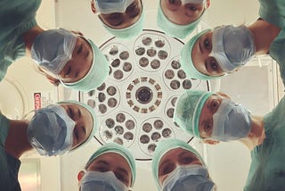 Doctors in scrubs and masks looking down at a patient. Menopause. Humor. Women’s Health. Aging.