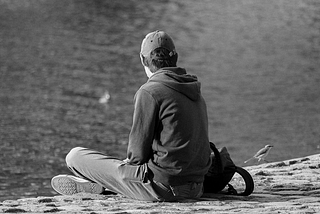 A grayscale photo of a man sitting near a body of water while looking at a bird.