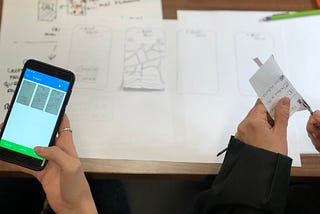 A hand holding a phone over some sketches. Another pair of hands cuts a piece of paper.