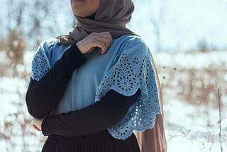 Girl with head scarf and blue bodice with her right arm diagonally across her chest reaching to her left shoulder. Her face is not visible.