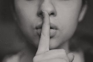 a grey-scale image of a person holding a finger to their lips, directing the viewer to be silent.