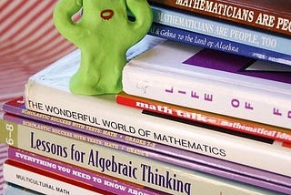 A green creature made of clay stands on a stack of math and logic books and appears to be distressed.