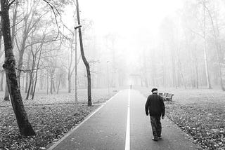 In a moody black-and-white photo, an old man shuffles along a lonely road.