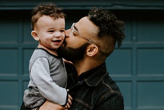 A young man with glasses and a beard holds his child on his shoulder and kisses him on the cheek. The child is smiling. They are standing in front of a green door.