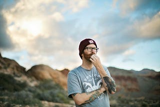 Image of a man with a beanie hat, glasses, and t-shirt pondering thoughts. Background are mountains and semi-cloudy sky, blurred out.