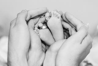 A black and white image of two sets of adult hands cupping an infant’s feet.