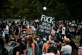 A man standing in a crowd protesting at a park holds up a black sign with white text that reads: Fuck special treatment for cops