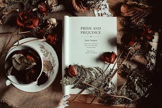 A book is opened to the title page, which reads “Pride and Prejudice, Jane Austen.” Dried roses are scattered over the book and a teacup and saucer sitting next to it.