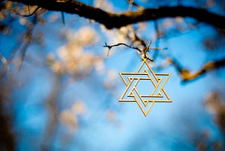 A gold star of david hangs from the branch of a tree against the background of a blue sky and white blossoms
