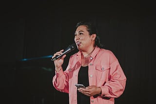 A woman against a black background in a business-casual outfit with a pale pink shirt speaks into a handheld microphone while holding her phone in her other hand.