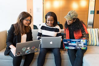 Three women sit on a couch in an office while using laptops.