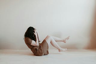 A woman sits on the floor, appearing angry as she lifts her legs and holds her head in her hands.