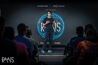 A photo of Nathan presenting, with the backdrop of the RAAIS logo.