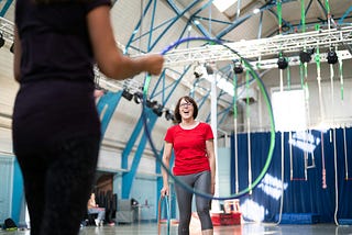 A woman in workout gear is laughing in front of a hula hoop held out by another woman.
