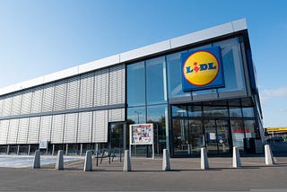 Modern Lidl supermarket with a glass facade, large Lidl logo, and accessible parking under a clear blue sky.