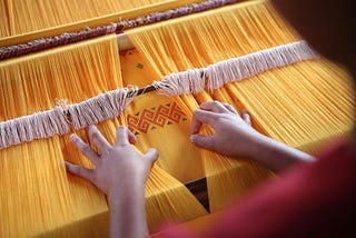 Hands weaving an orange fabric, parting the strands to reveal a geometric pattern in red