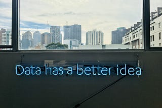 Neon Blue lettering. Phrase is ‘Data has a better idea’. It is below a window which has a view of a city skyline behind it.