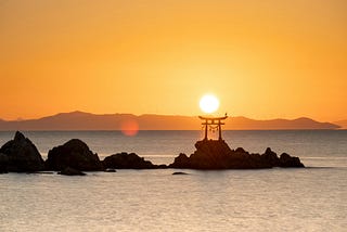 Sunrise over the sea with a silhouetted torii gate on a rock outcrop, a perched bird on the gate, faint hills and wind turbines on the horizon, under an orange sky.