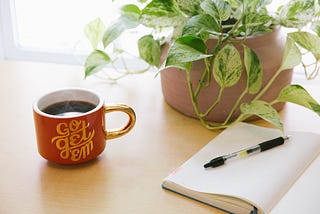 A coffee cup with Go get ’em written on it sits on a desk alongside an open notepad, a pen, and a plant