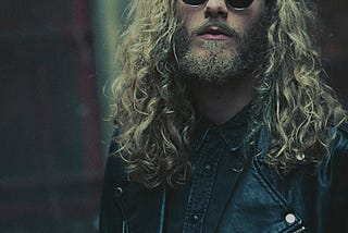 A young man with long curly hair stares directly at the camera. He is wearing dark glasses with round lenses and a black leather jacket.