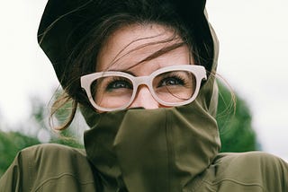 A woman wearing a black hat, large white glasses and wearing a jacket with the collar covering the bottom of her face.